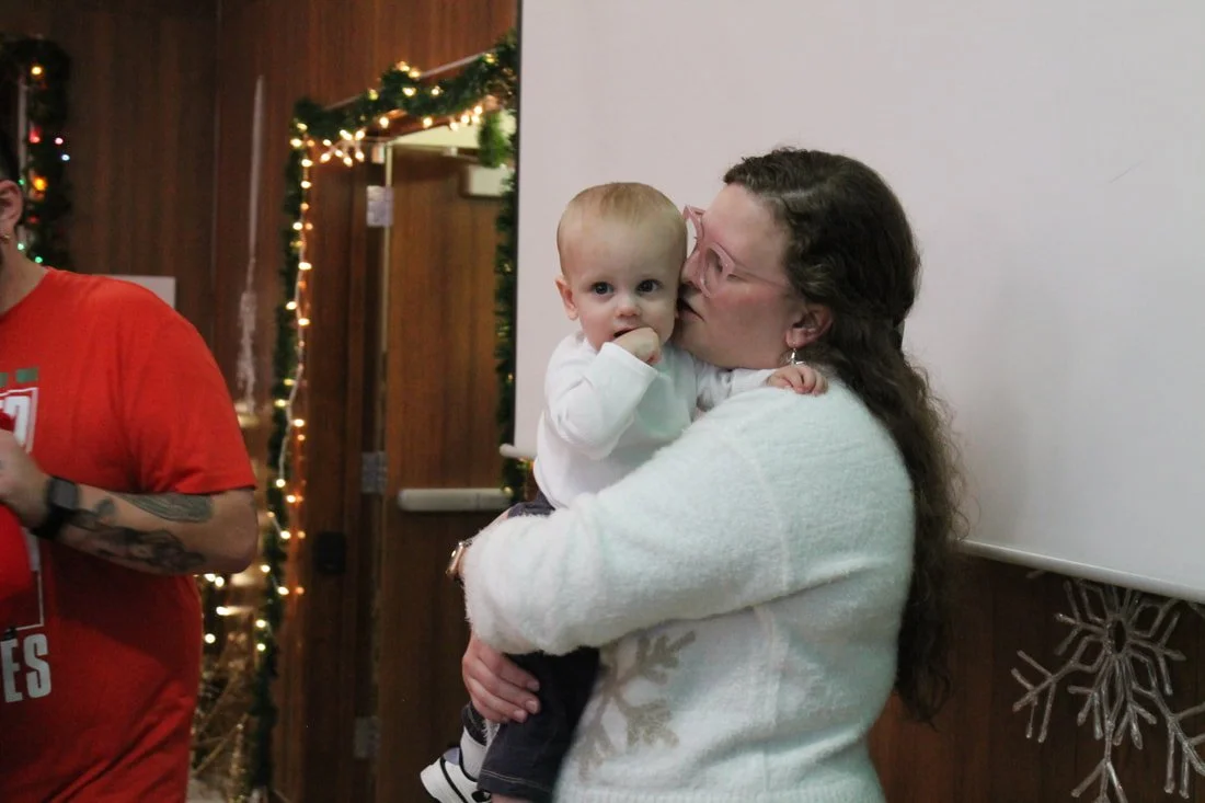 A woman holding a young boy at a holiday gathering, with Christmas decorations and festive lights in the background.