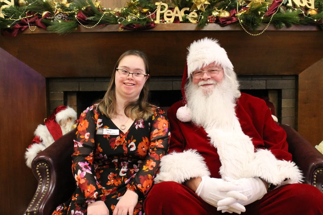 A young woman with glasses, wearing a floral dress, sitting next to a man dressed as Santa Claus in a red suit with a white beard, both smiling. They are sitting on a dark leather couch in front of a fireplace decorated with Christmas greenery, gold 