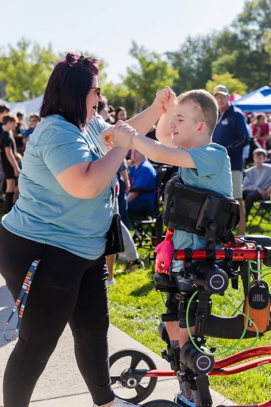 A woman and a boy in a wheelchair are dancing outdoors among a crowd at a park or festival, with trees and tents visible in the background.