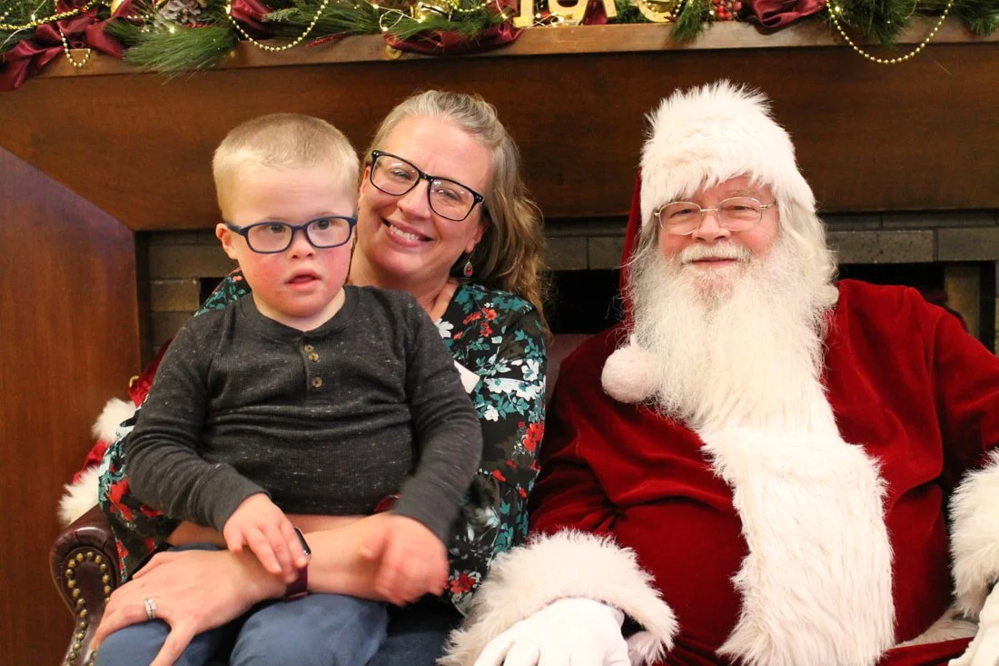 A young boy with glasses sitting on a woman's lap next to Santa Claus. They are in a festive setting with holiday decorations and garland in the background.
