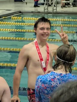 A young boy in a swimming pool wearing a red medal around his neck making a peace sign with his fingers, smiling, with a woman in a blue floral shirt in front of him.