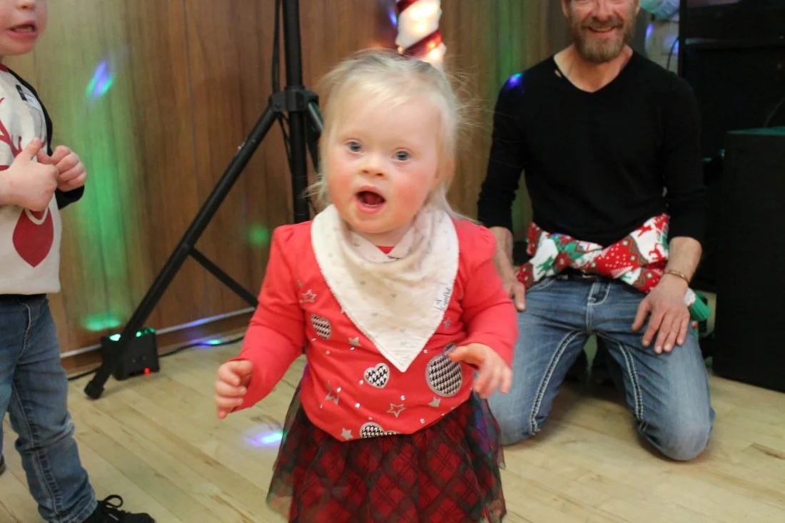 A young girl with blonde hair, wearing a red shirt, plaid skirt, and a white bandana, appears to be singing or talking with her mouth open at a holiday celebration. Behind her, a man kneels on the floor in a black shirt and holiday-themed apron, smil