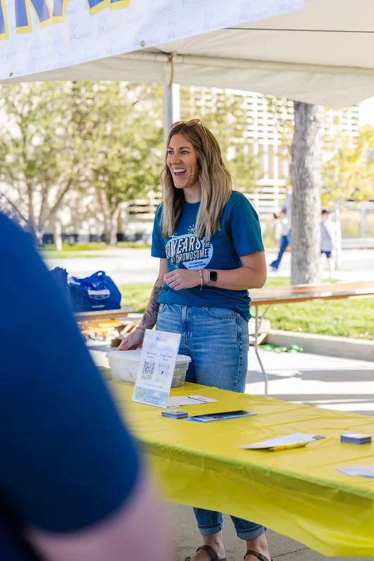 A woman with long blonde hair wearing a blue T-shirt and jeans, smiling and talking at an outdoor event booth. There is a yellow tablecloth on the table and a QR code stand.