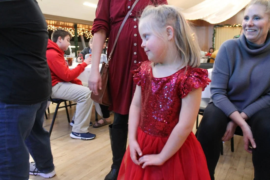 A young girl in a red, sequined dress stands indoors at a social gathering or party, surrounded by adults and other children.