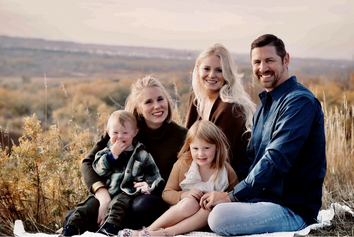 A family of five sitting outdoors in a field, smiling at the camera with rolling hills in the background.
