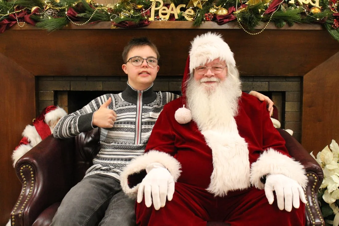 A young boy sitting next to Santa Claus, who is dressed in a traditional red suit with white fur trim, in front of a decorated Christmas mantle. The boy is giving a thumbs up and smiling, while Santa is smiling and sitting peacefully. The background 