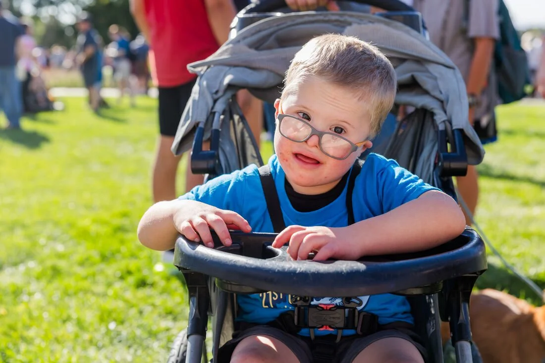 Young boy with glasses in a wheelchair outdoors, surrounded by people and green grass, smiling and looking at the camera.