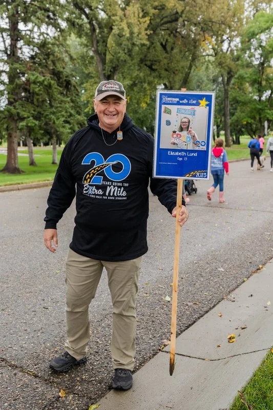 A man is standing on a park pathway holding a sign celebrating a young girl named Elizabeth Lund's 15th birthday at a community walk event in 2023. The man is wearing a black hoodie with a large blue '20' logo. In the background, people are walking i