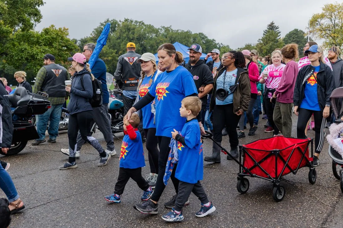 Group of people at an outdoor event, some wearing matching superhero T-shirts, walking on a wet pavement with motorcycles parked on the side and trees in the background.