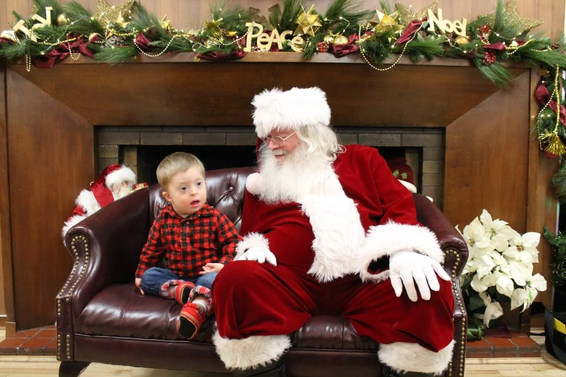 A young boy sitting next to Santa Claus on a bench, with a Christmas fireplace and holiday decorations in the background.