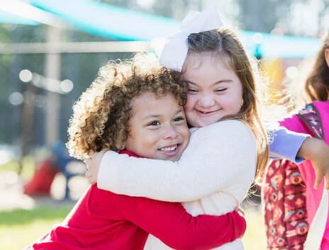Two young girls hugging and smiling outdoors on a sunny day.