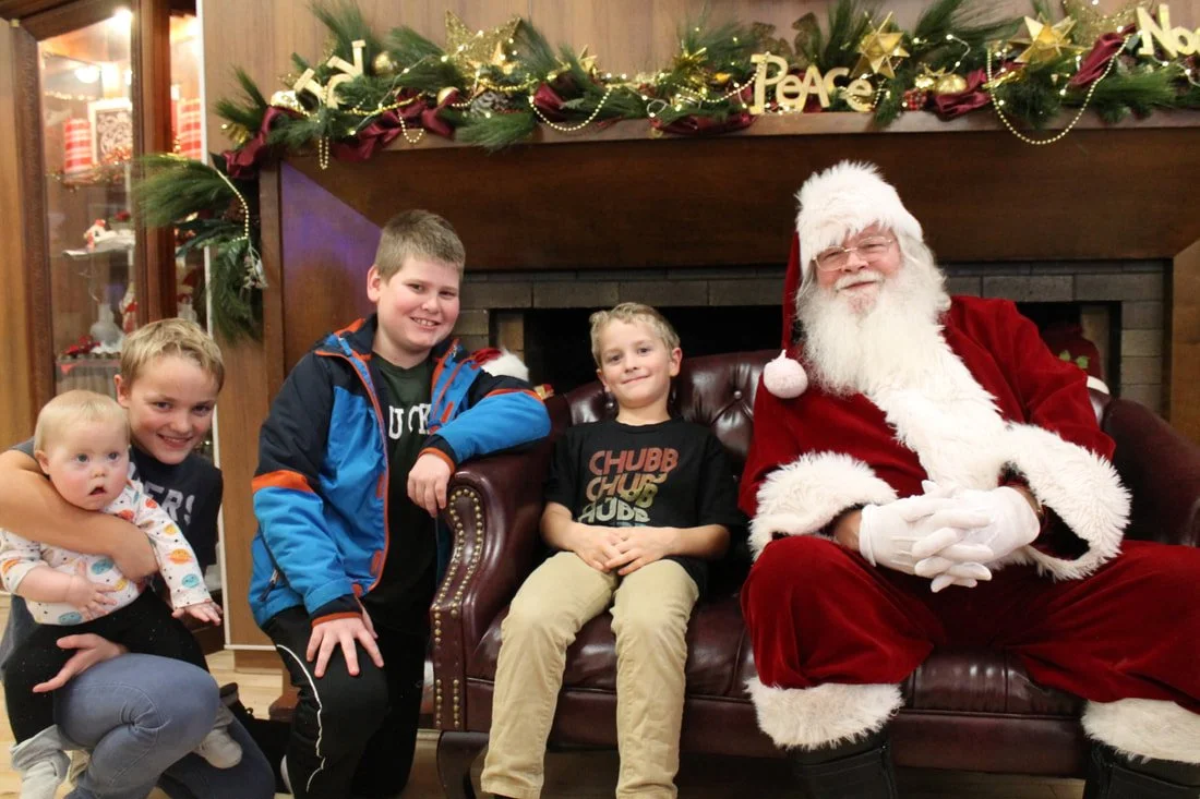 Four children sitting beside a man dressed as Santa Claus on a leather sofa in a room decorated for Christmas with greenery, gold ornaments, and Christmas signs overhead.