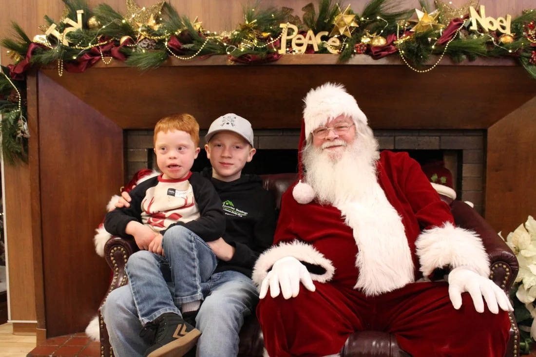 Two young boys sitting on Santa Claus's lap in front of a decorated fireplace with Christmas garland, ornaments, and holiday signs including 'Peace' and 'Noel'.