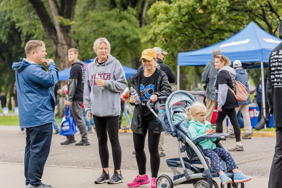People gathered outdoors at an event, including a man talking with a woman with blonde hair in a gray hoodie, a woman with a LA Dodgers cap holding a water bottle and pushing a stroller with a young girl, and other individuals near tents and trees.