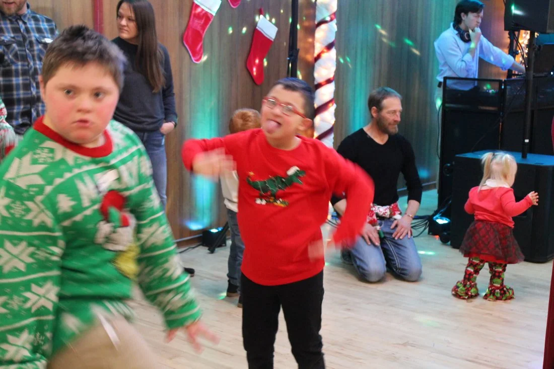 Children and adults dancing and celebrating at a Christmas party with holiday decorations, stockings, and a DJ in the background.