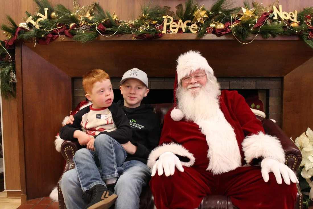 Two young boys sitting with Santa Claus on a decorated holiday backdrop. Santa is wearing a red suit and white beard, and the boys are dressed in casual clothing. The background features a Christmas garland with gold stars, ribbons, and holiday words
