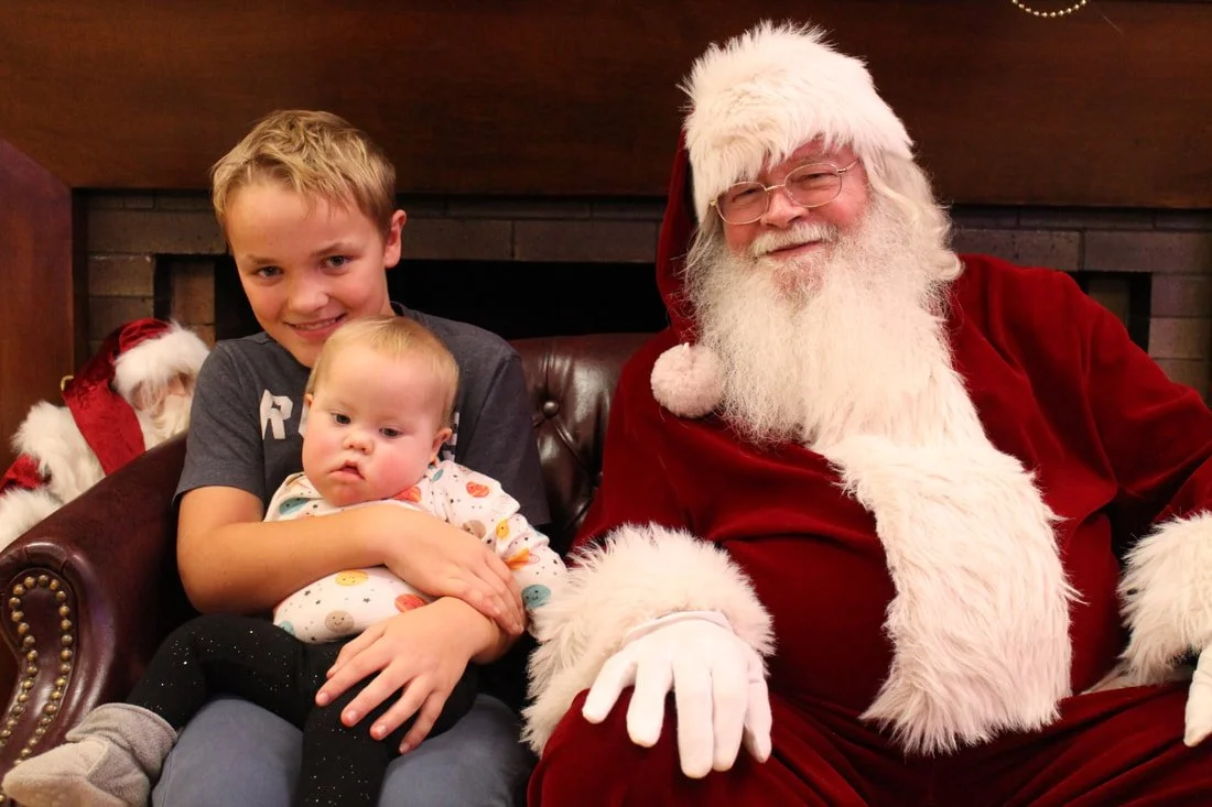 A young boy sitting on a Christmas-themed couch holding a baby girl, with Santa Claus sitting next to them, all smiling.