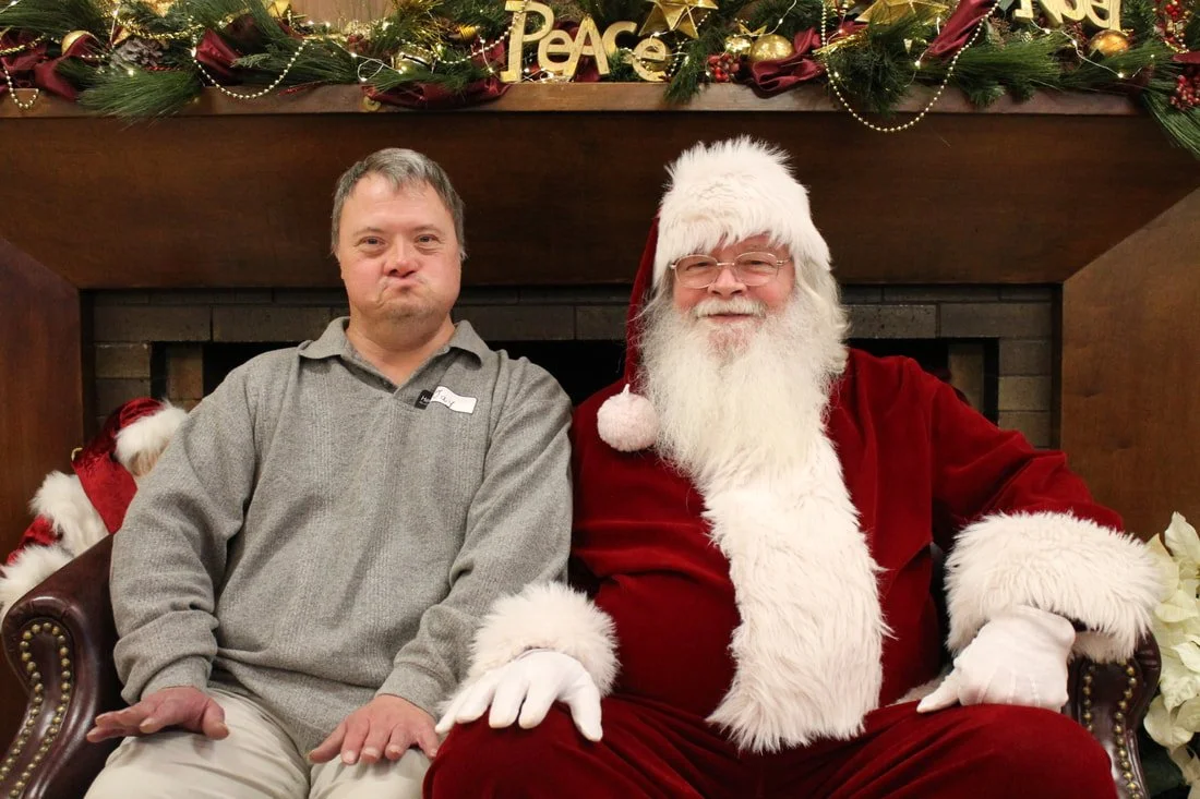 A man sitting next to Santa Claus, both seated on a brown leather bench in front of a decorated fireplace mantel with Christmas ornaments and greenery.