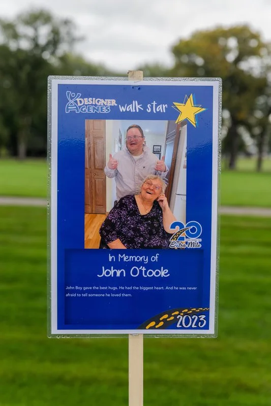 Memorial sign on a golf course in memory of John O'toole, featuring a photo of him with a woman, with text honoring his love and kindness, and the year 2023.