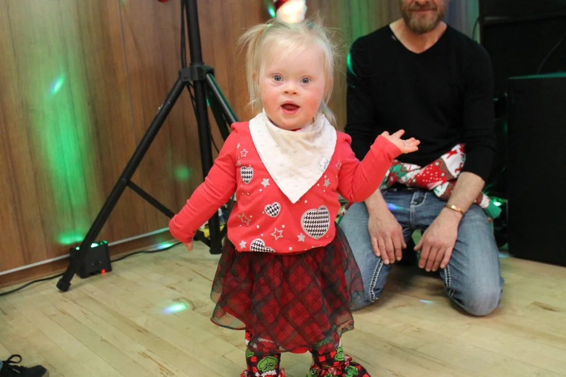 A young girl with blonde hair and a white bib, wearing a red Christmas sweater with black and white hearts and stars, a plaid skirt, and Christmas-themed tights, standing and smiling in a decorated indoor setting with a man kneeling behind her.