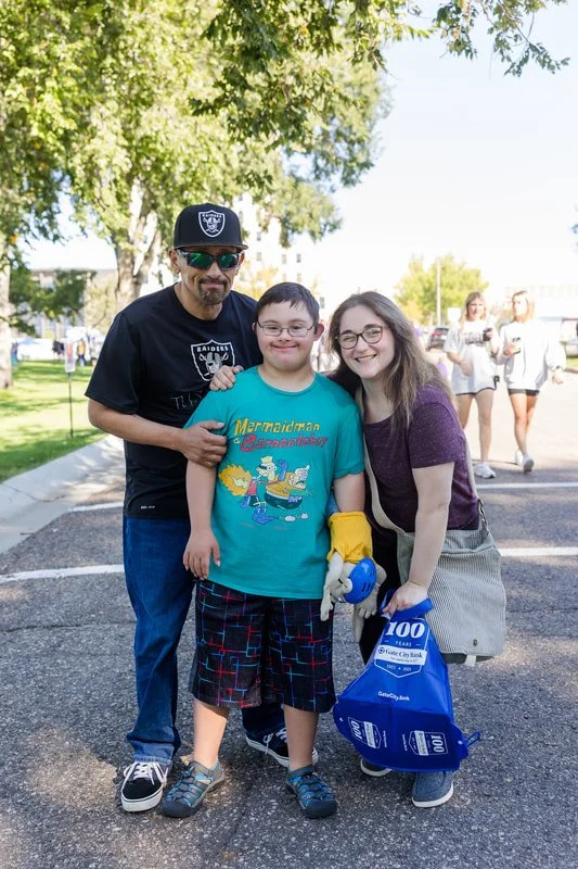A group of three people, including a man, a woman, and a boy, standing outdoors in a park-like setting with trees and a paved parking lot. They are smiling and posing for the photo, with the man and woman holding the boy's shoulders. The boy is weari