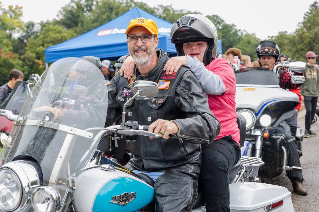A man and a young girl riding a Harley Davidson motorcycle at an outdoor event. The man is smiling and wearing a leather jacket and baseball cap, while the girl is wearing a helmet and pink hoodie.