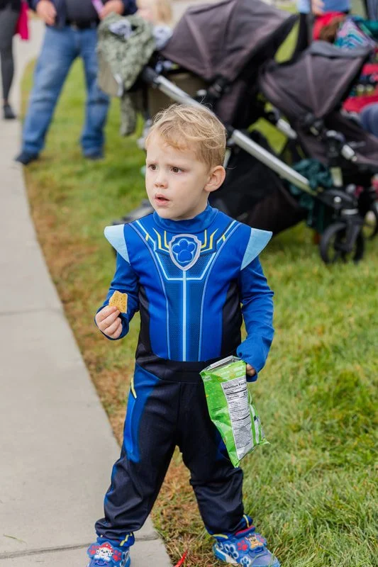 A young boy dressed in a Blue Power Rangers costume holding a chip and a bag of chips, standing on a sidewalk in a park with a stroller and people in the background.