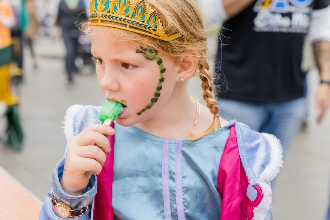 A young girl dressed as a princess and wearing a crown, with face paint and a snake design on her cheek, is eating a blue and green popsicle at an outdoor event.