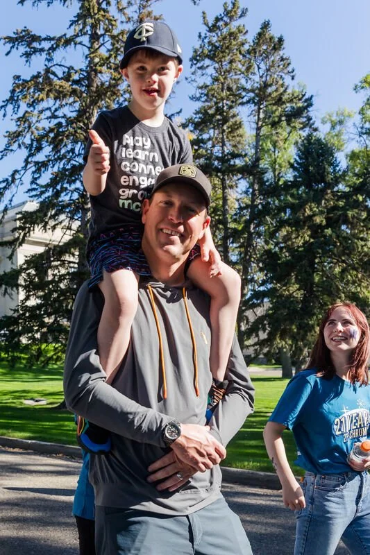 A man carrying a young boy on his shoulders, both smiling, outdoors on a sunny day with trees in the background. The boy is giving a thumbs-up.