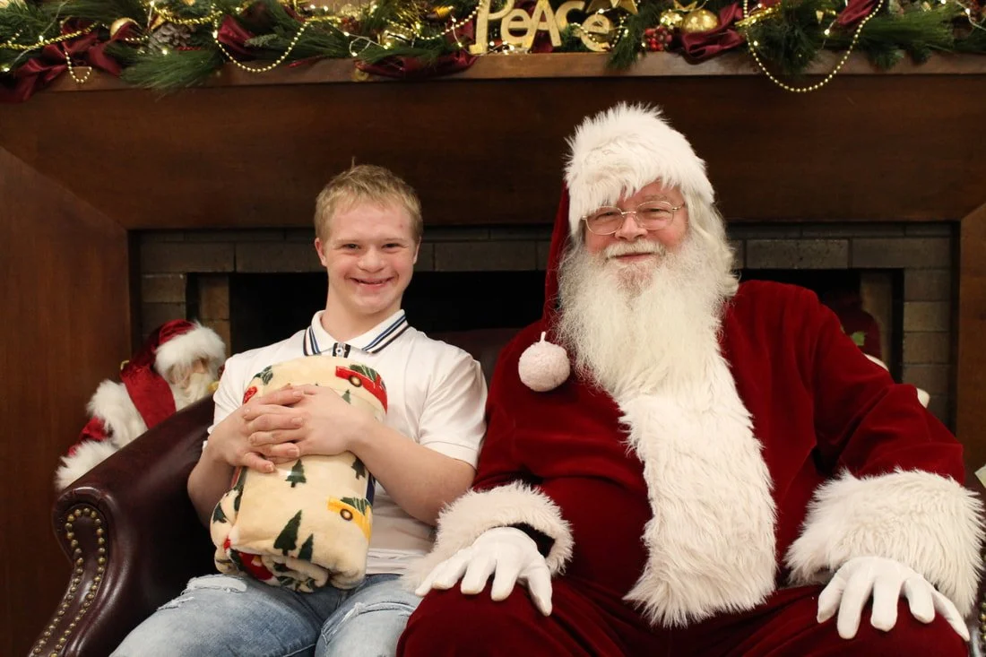 A young boy sitting next to Santa Claus, holding a wrapped gift. They are in front of a decorated fireplace with Christmas ornaments and garland, with the word 'PEACE' displayed on the mantle.