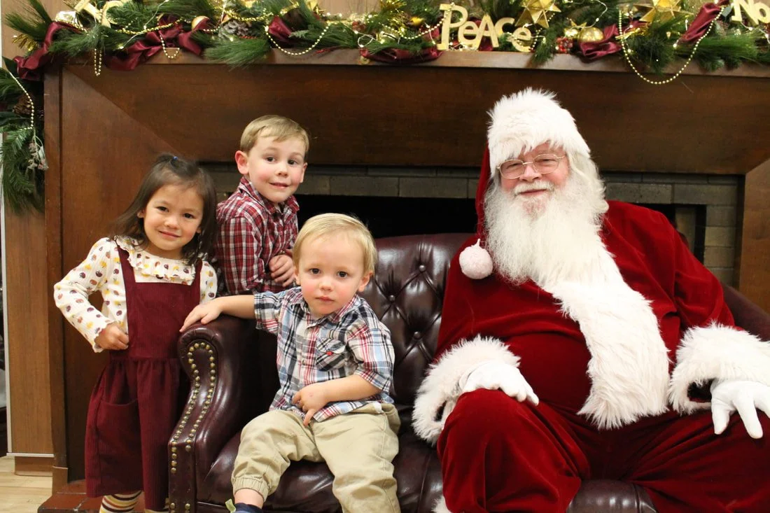 Three children and Santa Claus in a festive setting with Christmas decorations on the mantel, including greenery, ornaments, and a 'Peace' sign, sitting on a leather couch.