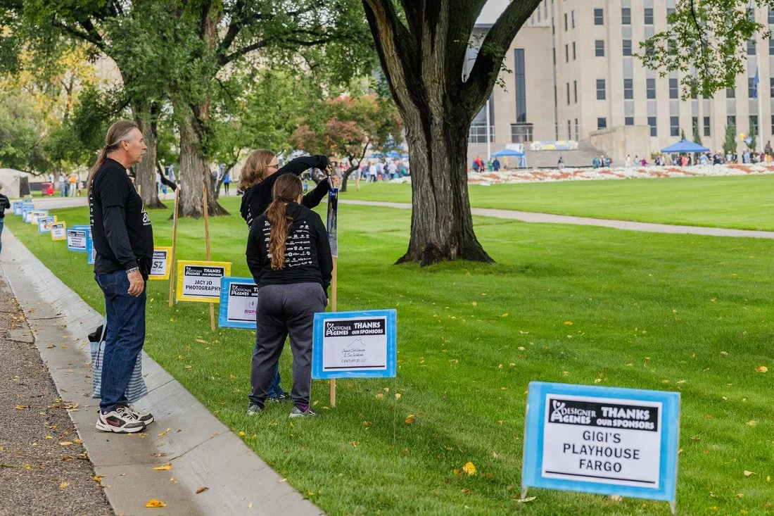 Group of four people, three women and one man, standing on a sidewalk and looking at signs in a park with trees and grass. The signs are thank you messages from sponsors. The background shows a building and some tents.