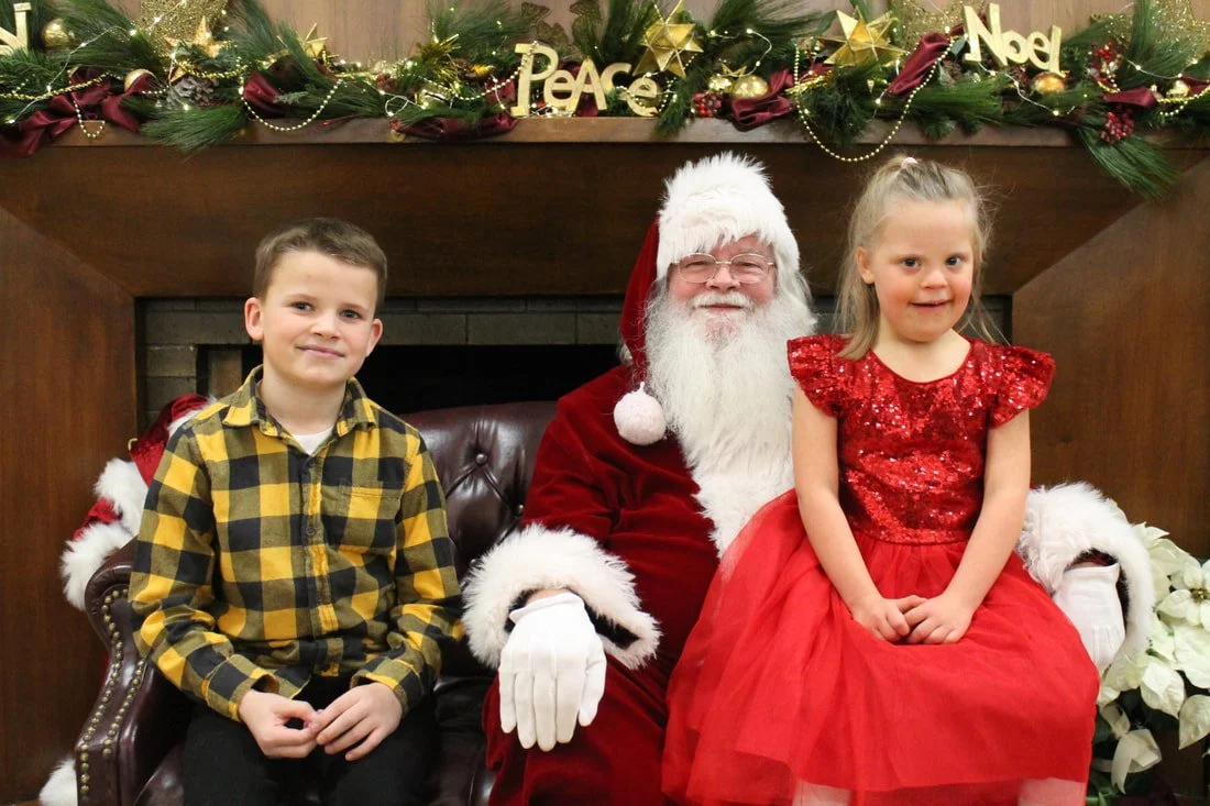 A boy and a girl sitting with Santa Claus in front of a Christmas fireplace decoration with greenery, red ribbons, gold ornaments, and 'Peace' and 'Noel' signs.