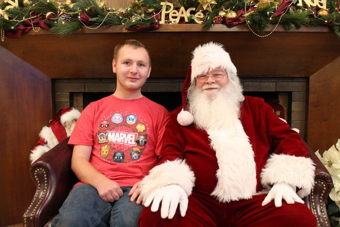 A young man sitting next to Santa Claus on a decorated holiday couch in front of a fireplace, with Christmas garland and lights above.