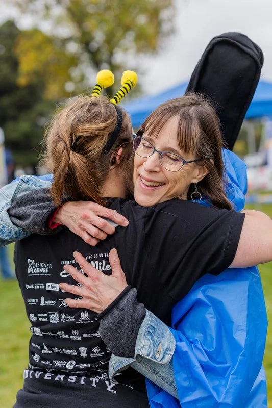 Two women hug smiling at an outdoor event, one wearing bee antenna headband and the other wearing glasses and a blue rain jacket.