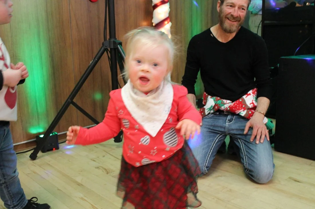 Young girl dancing at a holiday party, with a man in the background smiling, decorated with Christmas lights and garland.
