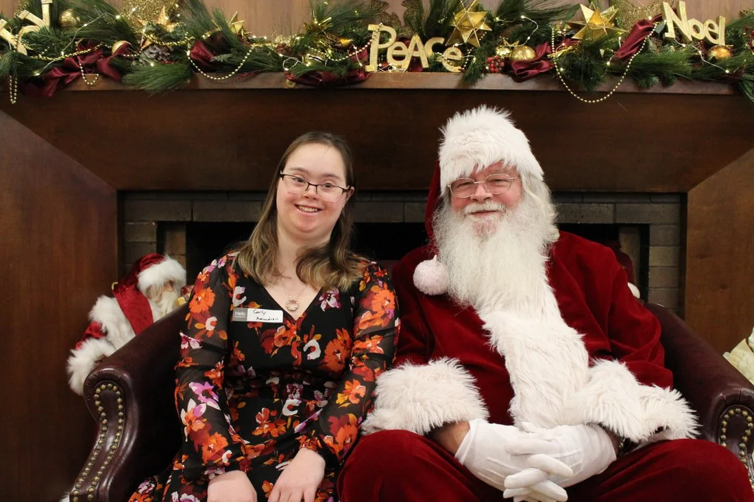 Woman and Santa Claus sitting in front of a decorated fireplace with Christmas garland, lights, and ornaments.