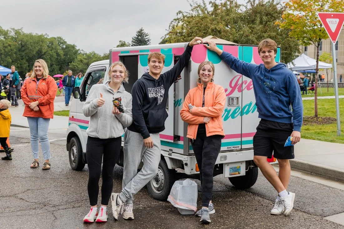Group of four young people posing in front of an ice cream truck at an outdoor event, with a woman and child in the background and several tents and trees.