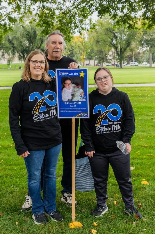 Three people standing outdoors in a park, holding a sign with a photo of a young girl. They are wearing matching black hoodies celebrating 20 years of Extra Mile charity. The sign features the girl, named Sakuara Pretti, with her age, 25, and the yea