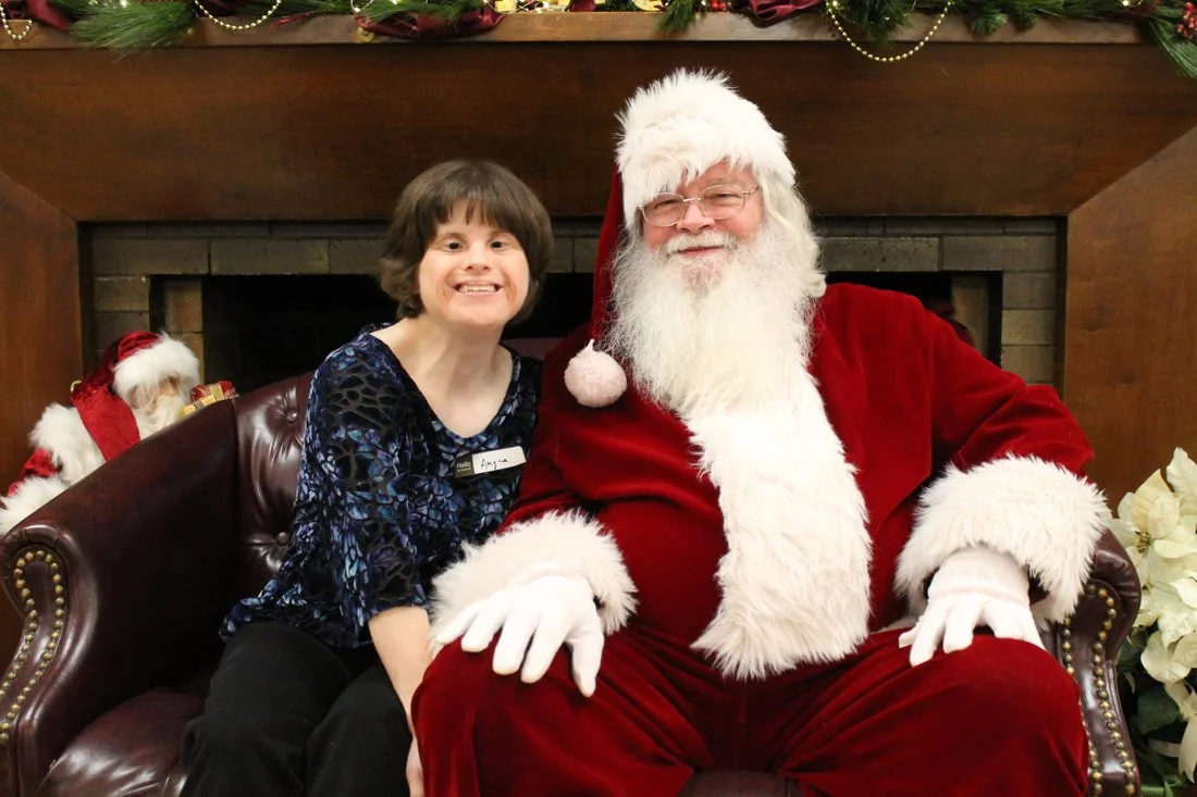 A young child with short brown hair and a wide smile sitting next to Santa Claus, who is wearing a red suit, glasses, and a white beard. They are sitting on a leather bench in front of a fireplace decorated with holiday garland and white flowers.