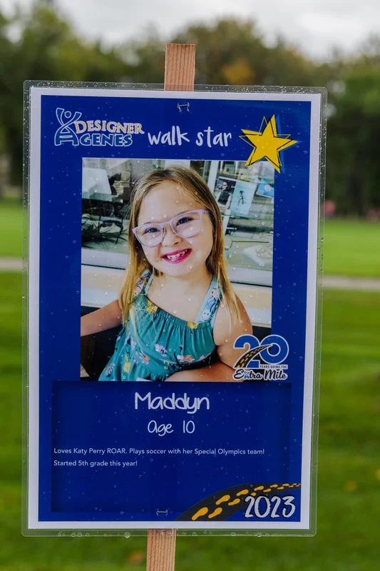 A young girl named Maddyn, age 10, is part of a walk star event for Designer Genes, celebrating 20 years in 2023. She is dressed in a teal dress with floral patterns, wearing glasses, and smiling at the camera.