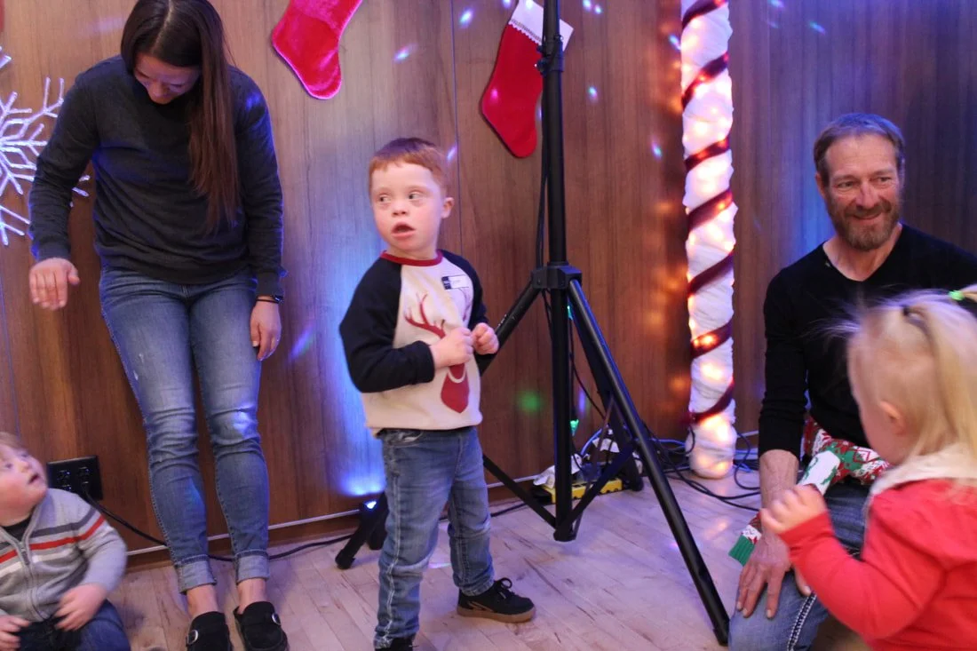 People celebrating Christmas indoors, with a wooden wall decorated with Christmas stockings, snowflake lights, and striped candy cane poles, during a festive gathering.