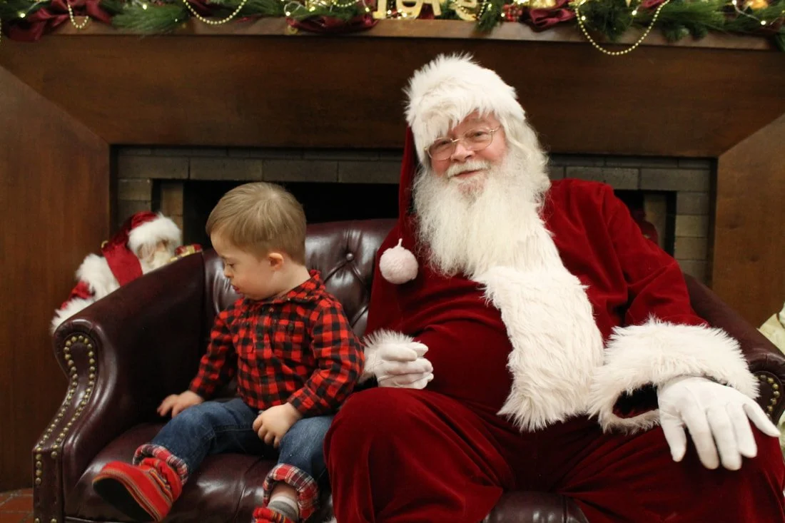 A young boy sitting on Santa Claus's lap at a Christmas event, with festive decorations in the background, including a decorated mantle with garland and lights.