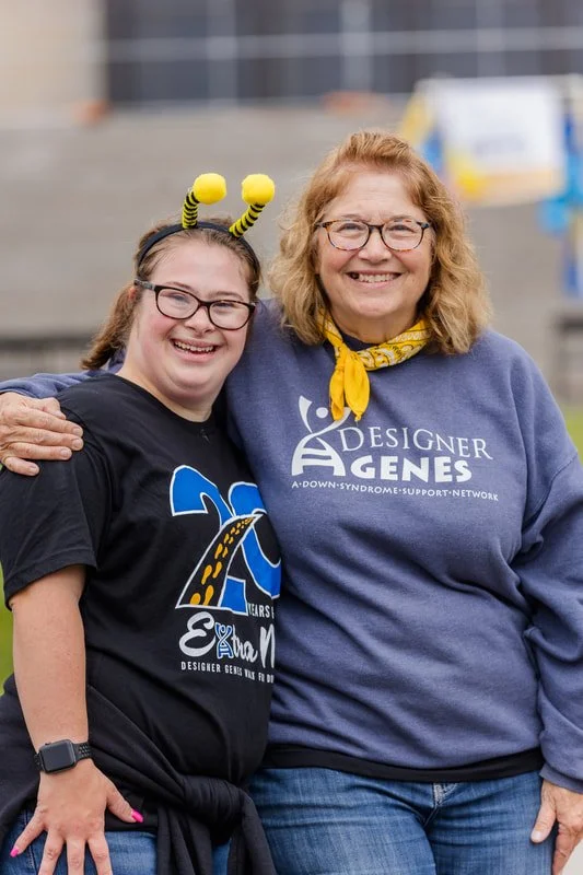 Two women smiling and standing outdoors with their arms around each other. The woman on the left is wearing a black T-shirt with a wheelchair logo and a yellow headband with antennae, while the woman on the right is wearing glasses, a blue sweatshirt
