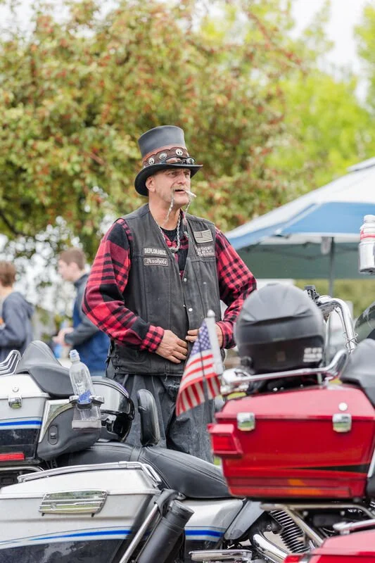 A man wearing a black top hat, plaid shirt, and vest standing next to motorcycles at an outdoor event.