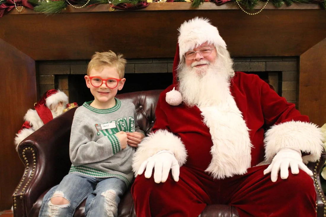 A young boy with glasses and a gray sweater sitting next to Santa Claus in a red suit, sitting on a leather armchair in front of a fireplace decorated with Christmas garland and ornaments.