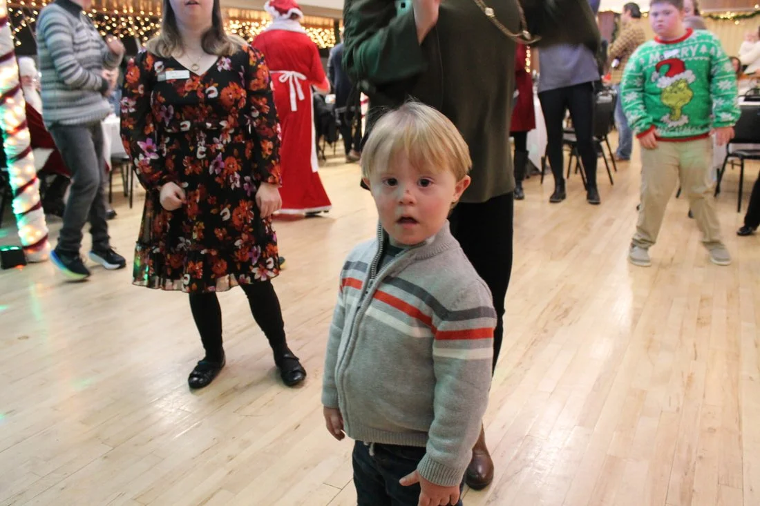 Young boy with blonde hair wearing a gray sweater with colorful stripes stands on a wooden floor at a holiday party, surrounded by other people in seasonal attire.