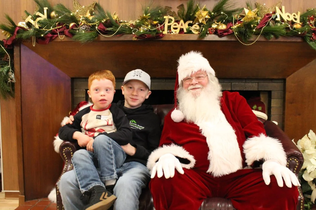 Two young boys sitting with Santa Claus in front of a decorated fireplace mantle with Christmas ornaments and greenery.