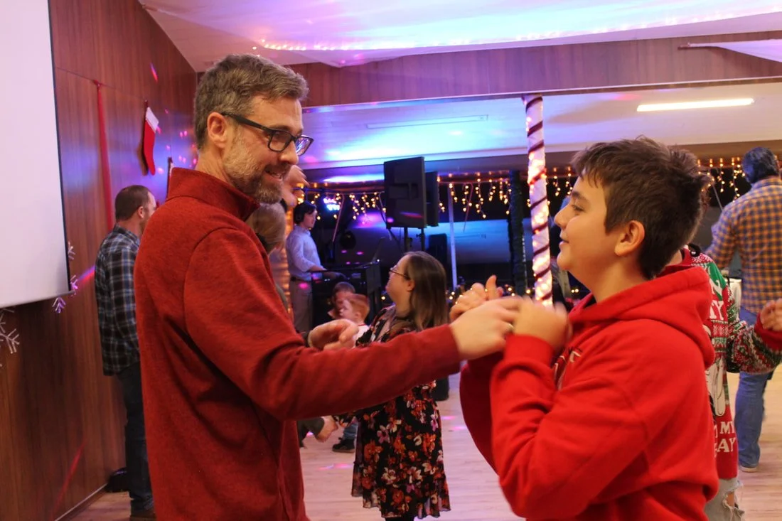 A man and a boy holding hands and dancing at a holiday party with Christmas decorations and string lights in the background.