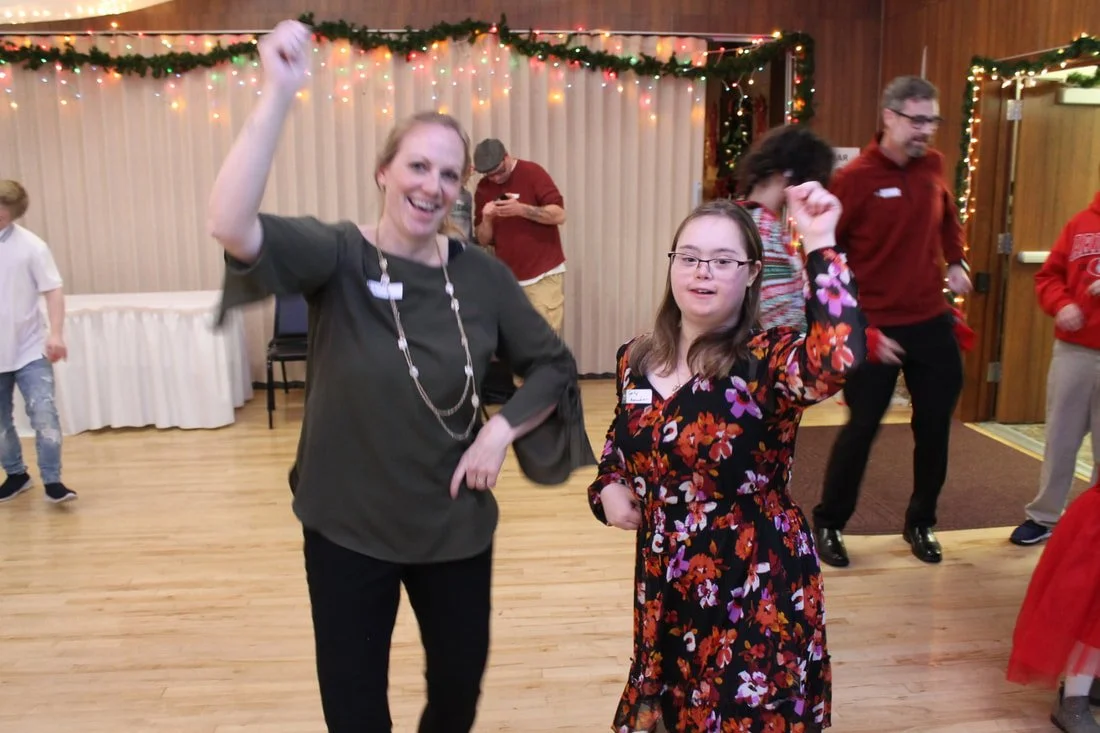 Two women dancing and celebrating at a holiday party with string lights and greenery decorations in the background.